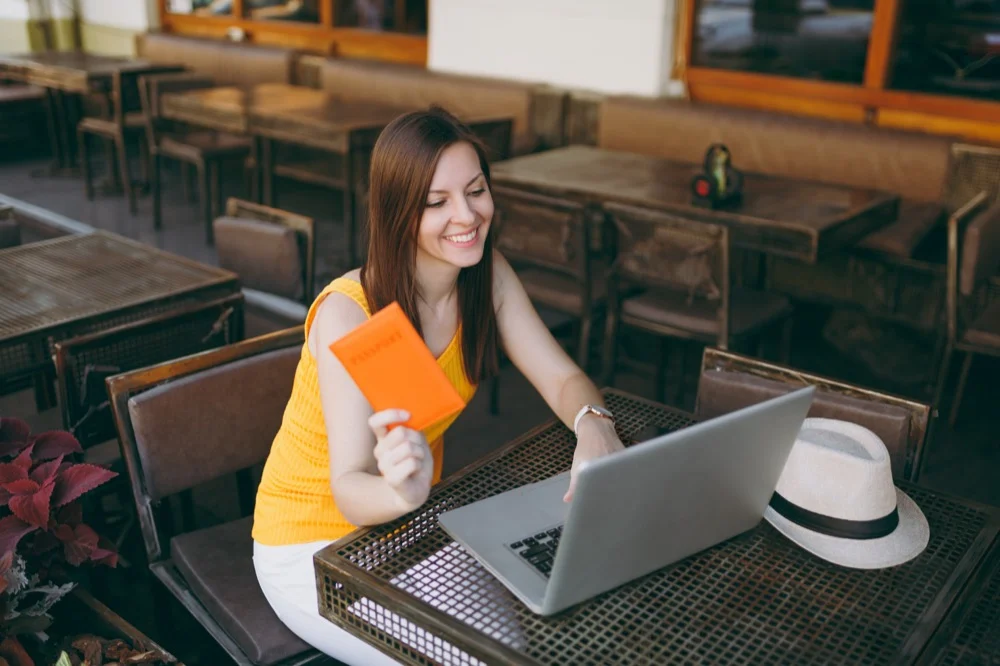 A smiling woman sitting at an outdoor cafe holding a passport while using a laptop with a travel hat on the table