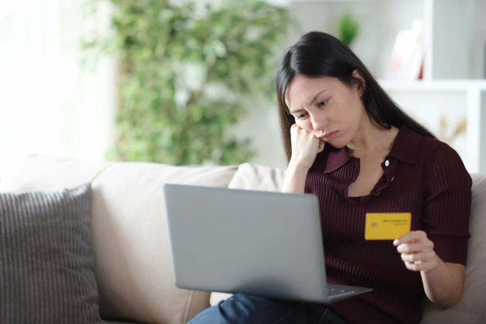 Worried woman holding a credit card while looking at a laptop on a sofa