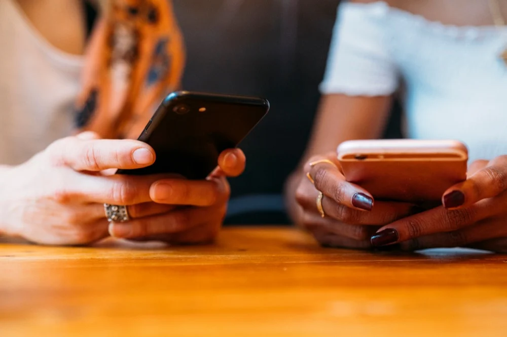 A close-up of two people's hands using smartphones while sitting at a wooden table