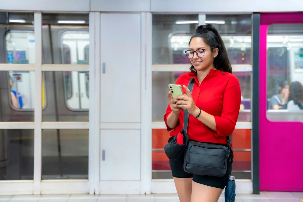 A smiling woman in a red shirt and glasses looking at her phone while standing in front of subway platform doors