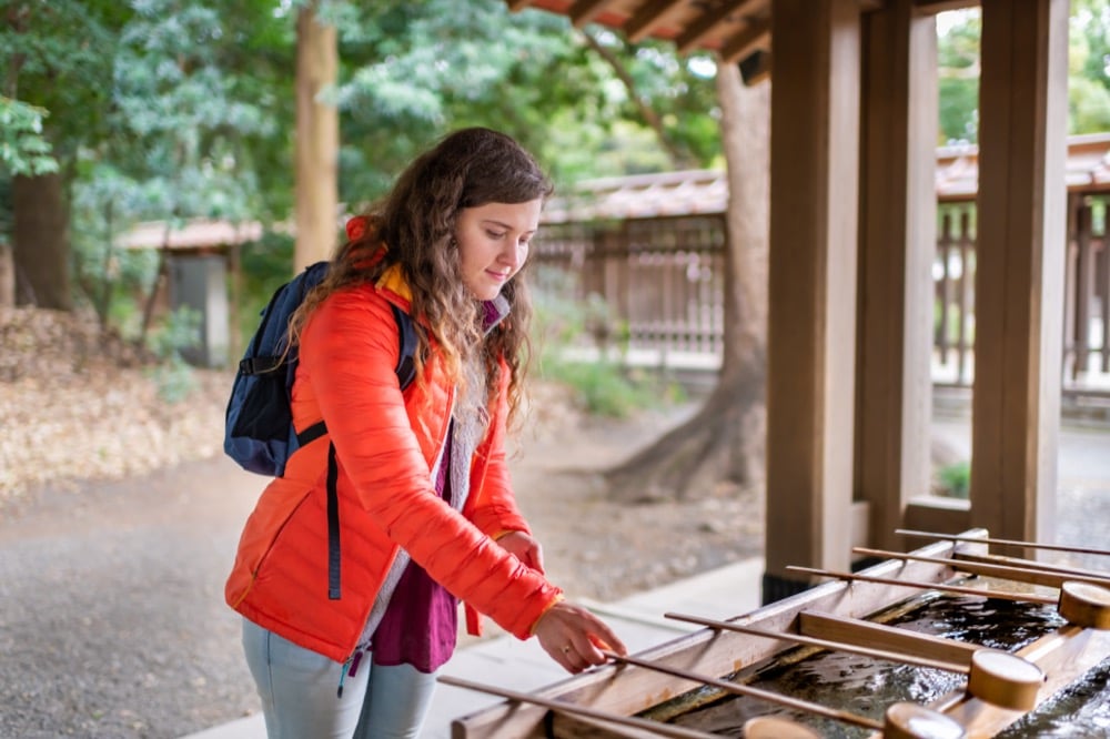 Female tourist in orange jacket performing purification ritual at Japanese Shinto shrine temizuya