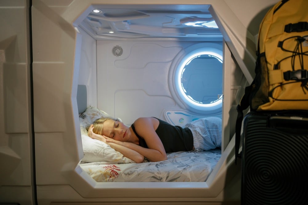 A female traveler sleeping peacefully inside a modern Japanese capsule hotel pod with blue ambient lighting
