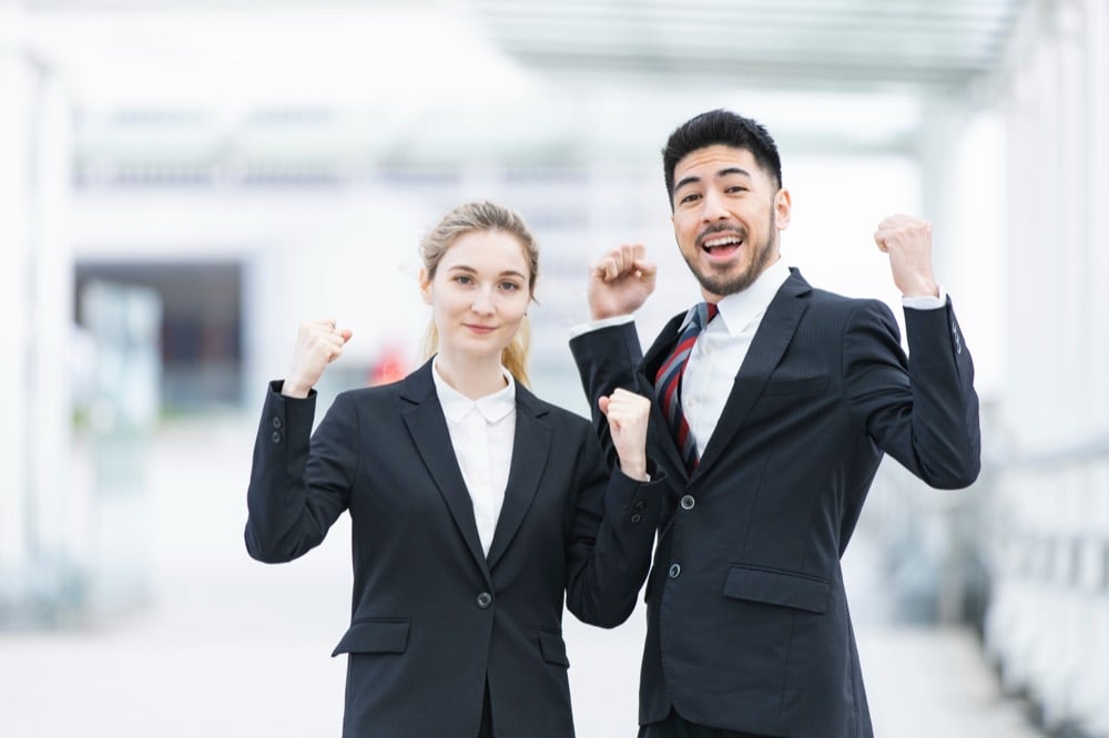 Two business professionals in black suits celebrate with raised fists and big smiles outdoors