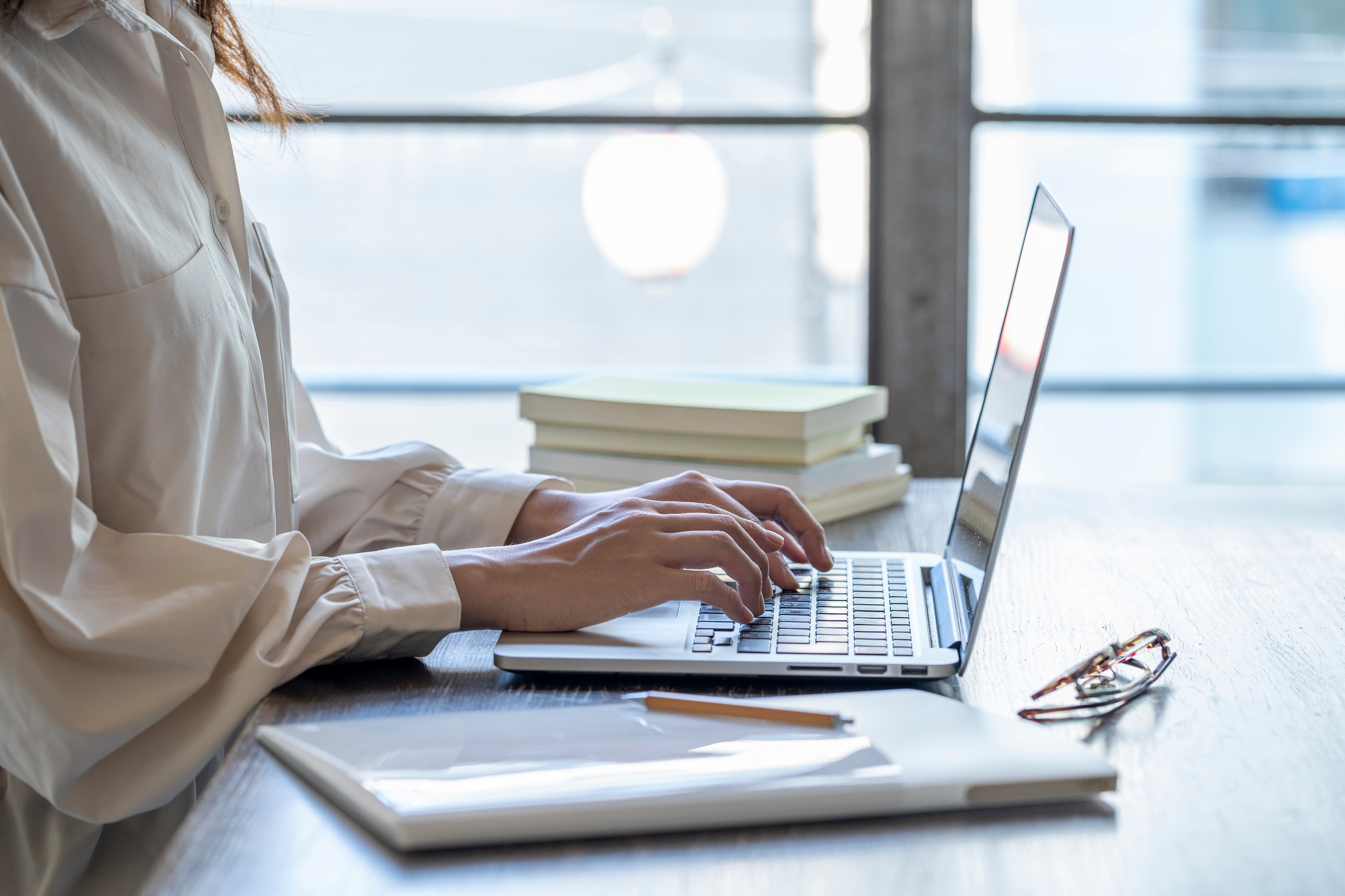 Woman in white blouse typing on a laptop at a desk with books and glasses nearby