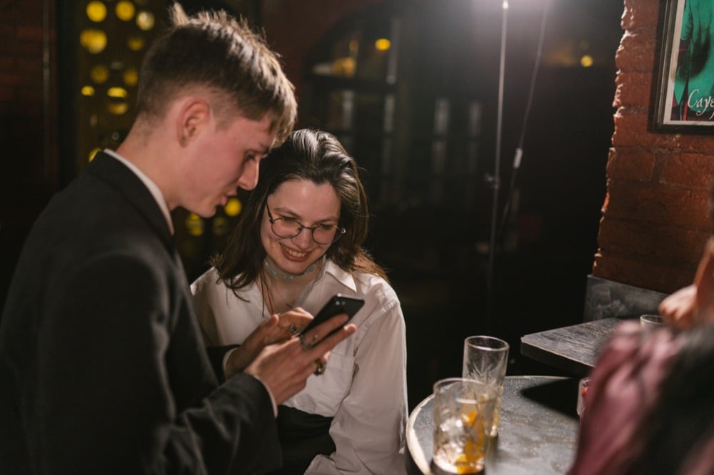 Two young people smiling at a smartphone screen at a bar