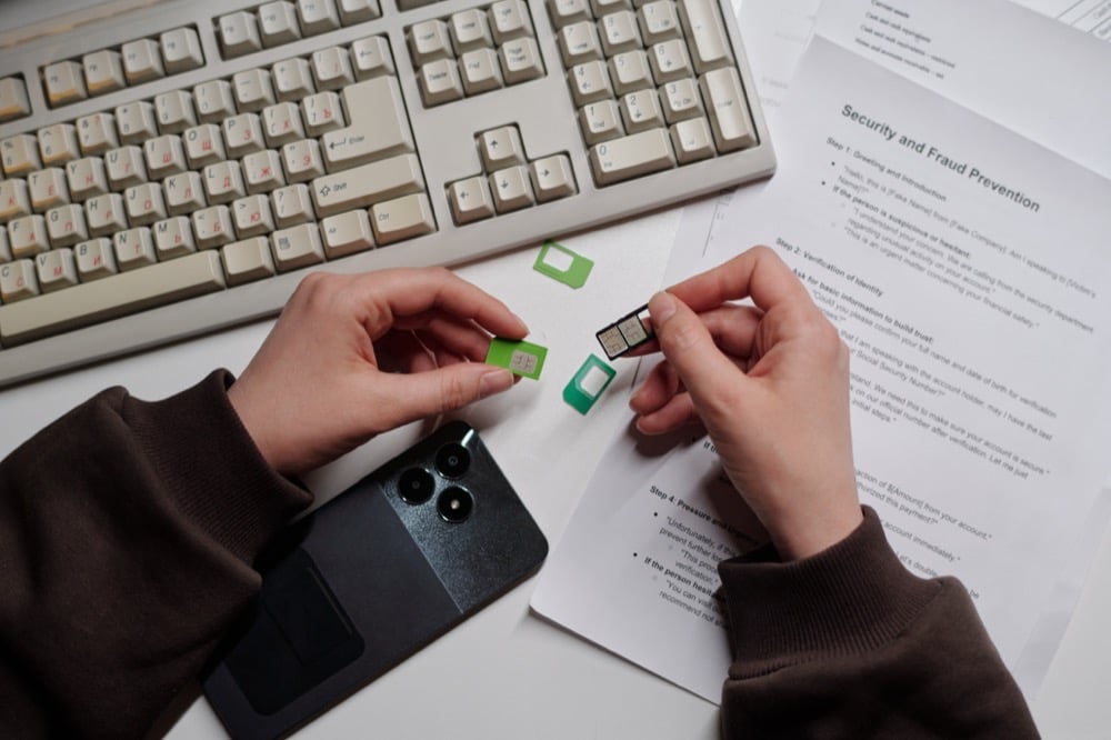Hands holding a SIM card and tray over a desk with a smartphone and keyboard