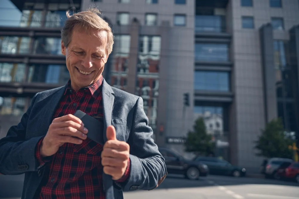 A smiling middle-aged man giving a thumbs up while using a smartphone on a city street