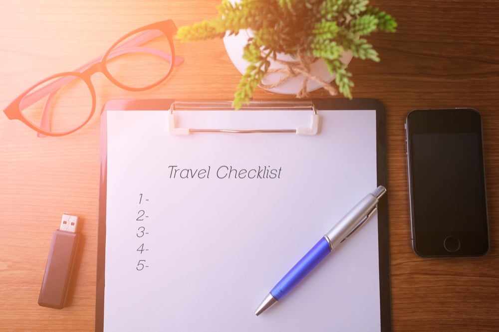 A travel checklist on clipboard with pen, glasses, and a smartphone