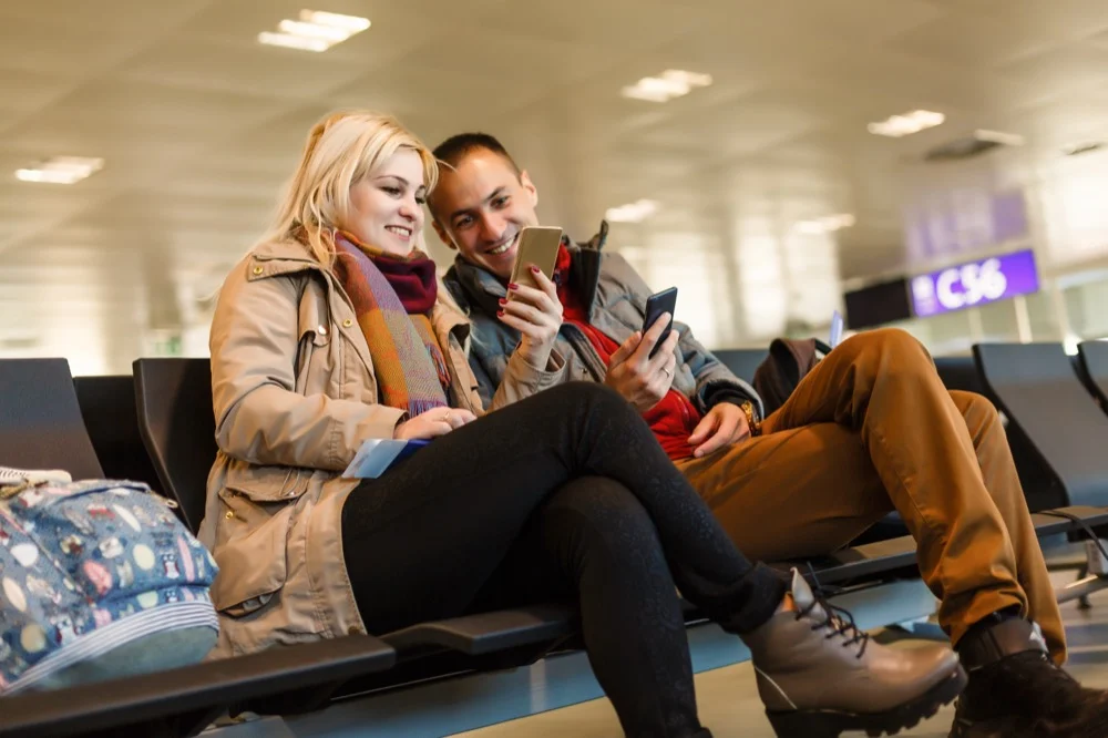 A smiling couple sitting together at an airport terminal and looking at a smartphone