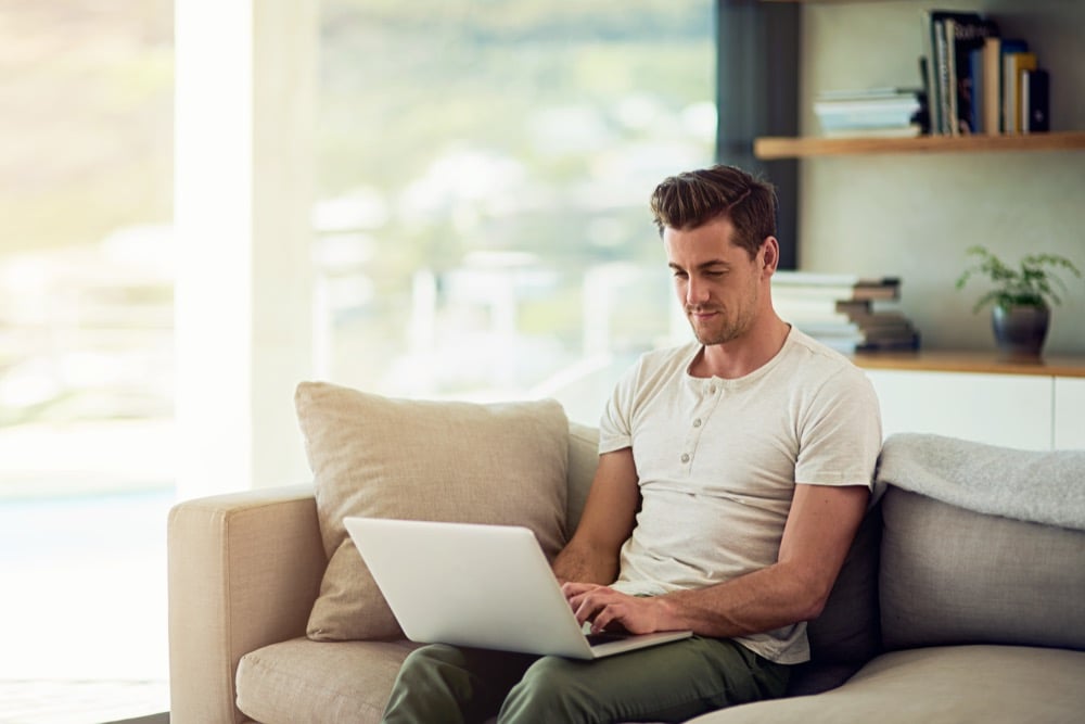 A smiling young man works on a laptop while sitting on a sofa in a bright living room