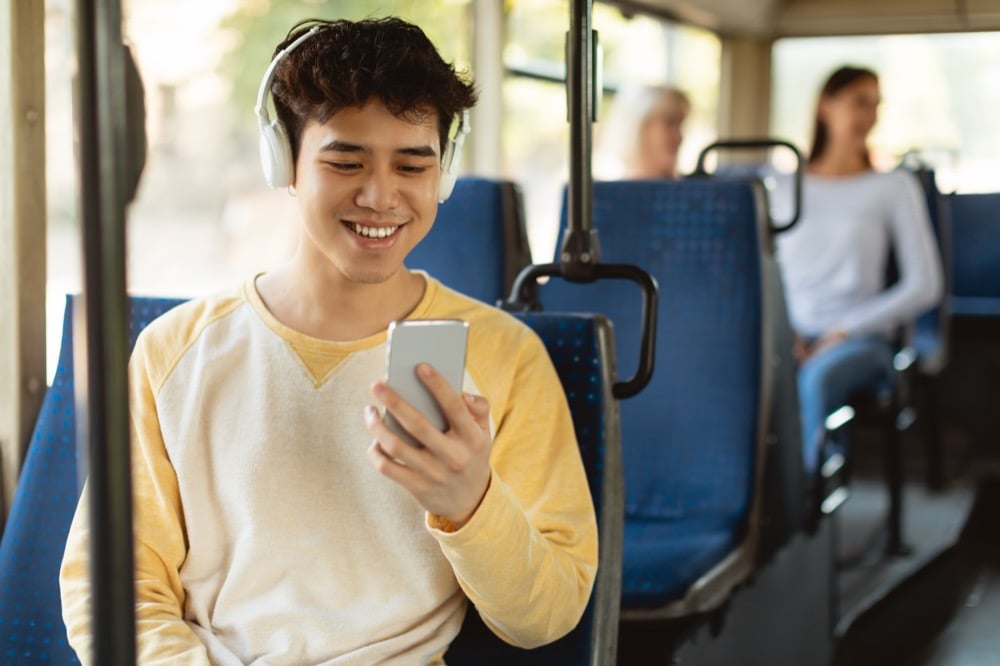 A smiling young man wearing headphones using a smartphone while riding a public bus