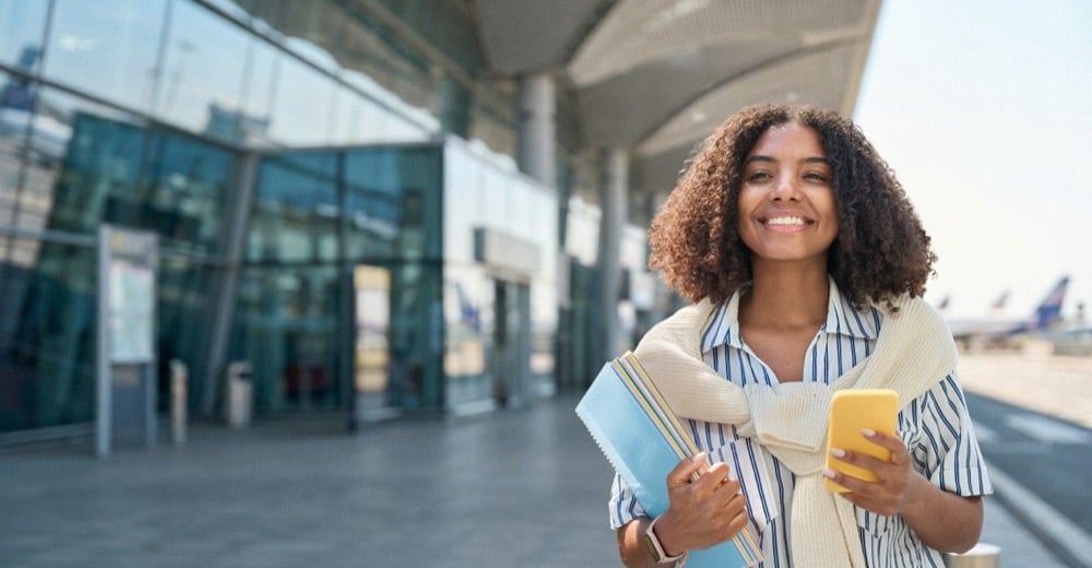 A smiling woman holding a smartphone and passport outside a modern airport terminal entrance