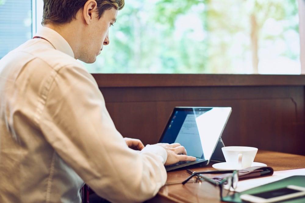 A man in a white shirt types on a laptop at a cafe table with a coffee cup nearby