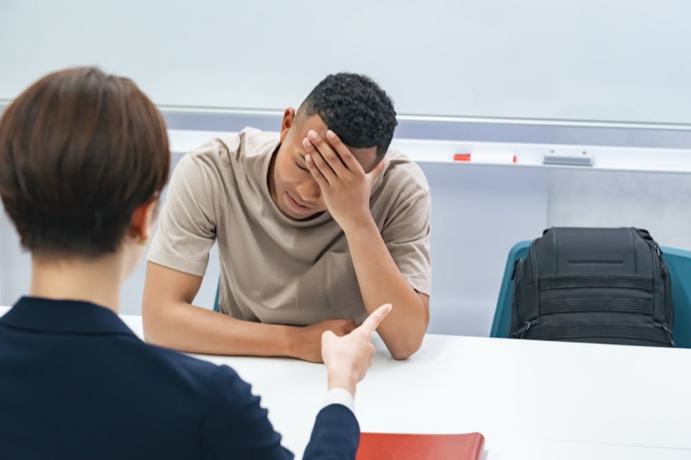 A young man holds his head in distress at a desk while another person points at him