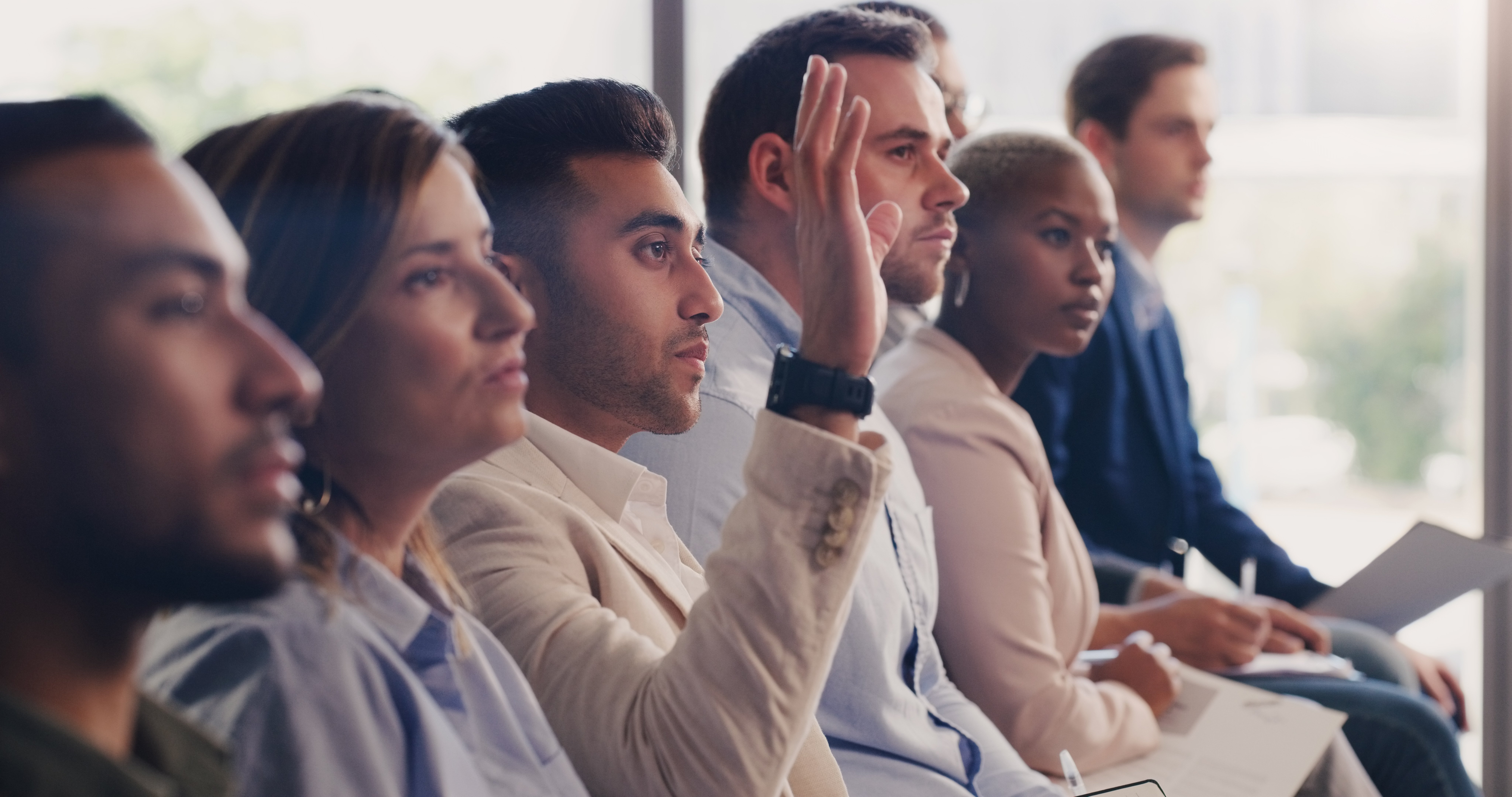 Diverse group of professionals sitting in a row at a seminar, one raising hand