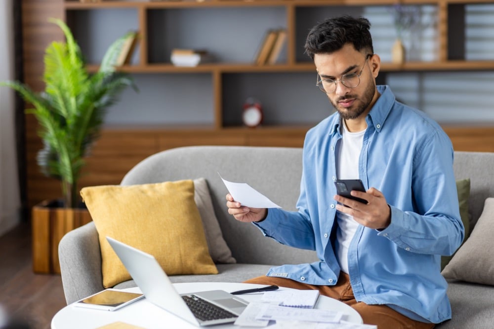 A young man reviewing a document and smartphone while working at home with a laptop