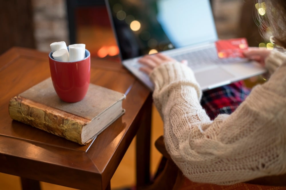 A person online shopping by a fireplace with hot cocoa and books nearby