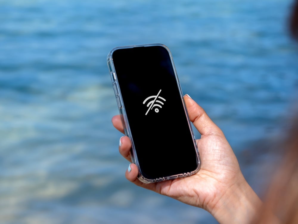 A hand holding smartphone with no Wi-Fi icon over blue water