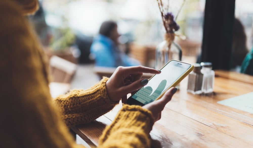 Woman in yellow knit sweater typing on a smartphone at a cafe table