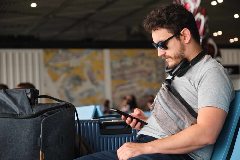 A man with sunglasses and headphones sitting at an airport and looking at his smartphone