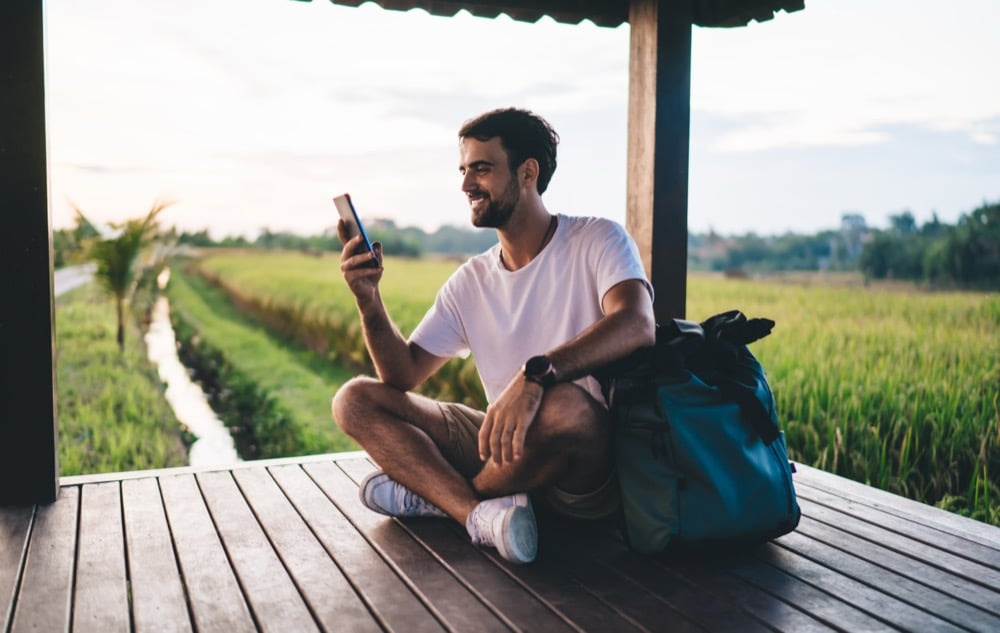 A smiling traveler with a backpack using a smartphone outdoors in a rural landscape