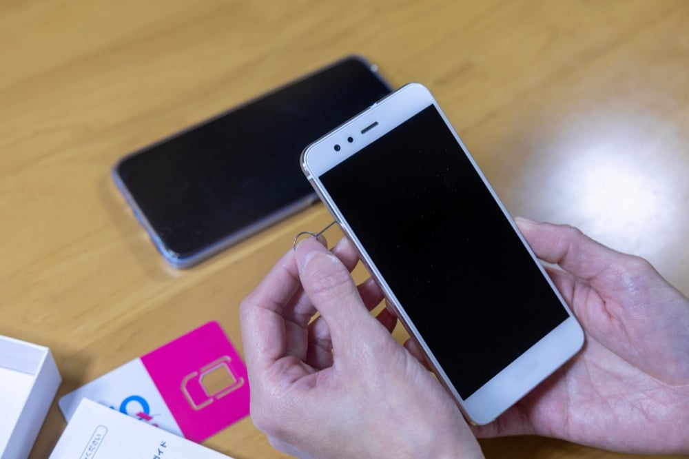 A person uses a SIM ejector tool to open a smartphone's SIM tray, with a pink SIM card package nearby