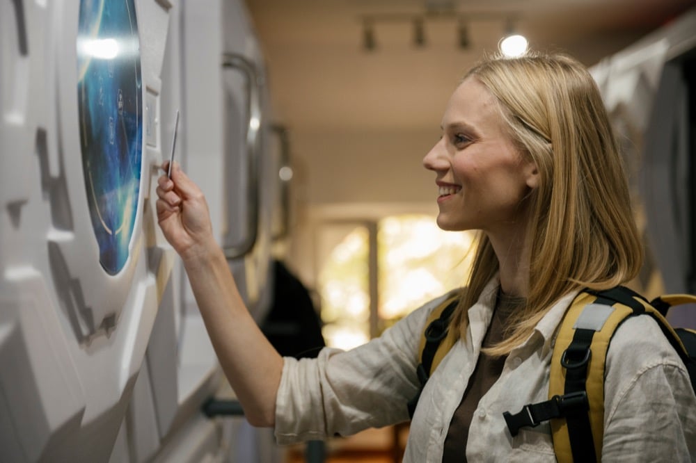 A smiling blonde woman with a yellow backpack using a key card to access a capsule hotel room