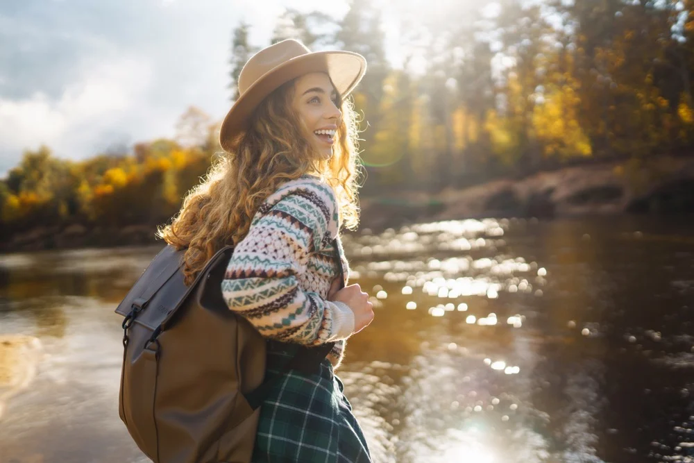 Smiling female traveler with a backpack and hat standing by a sunlit autumn river in nature