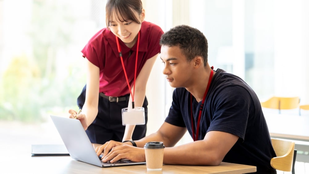 Two colleagues with red lanyards collaborate at a laptop as a smiling woman leans over while a man types