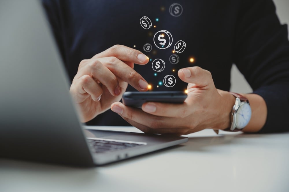 A person using a smartphone with floating dollar coin icons representing mobile payment or digital finance