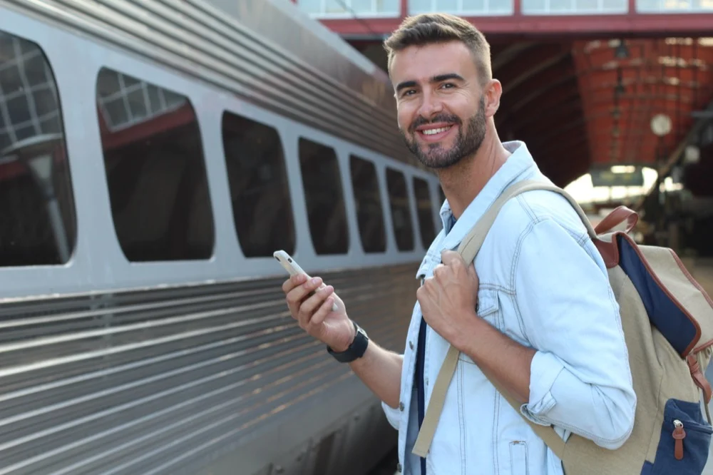 A smiling traveler with a backpack using a smartphone at a train station