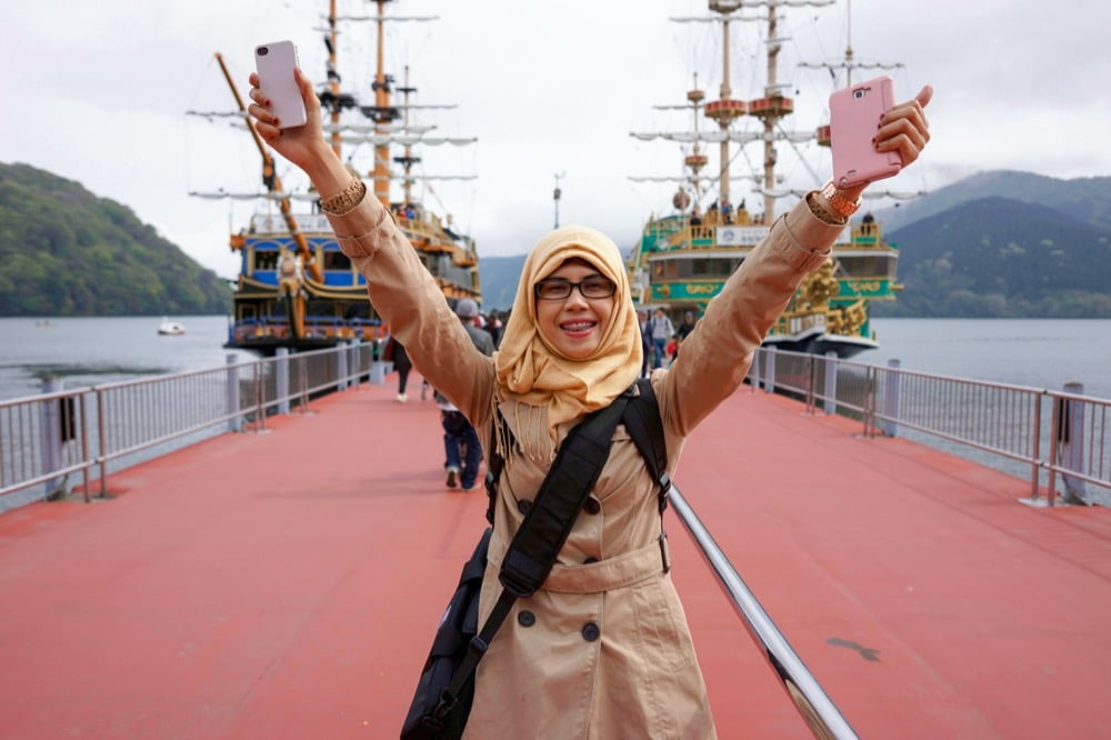 A smiling woman in a hijab raising two smartphones in front of a ship