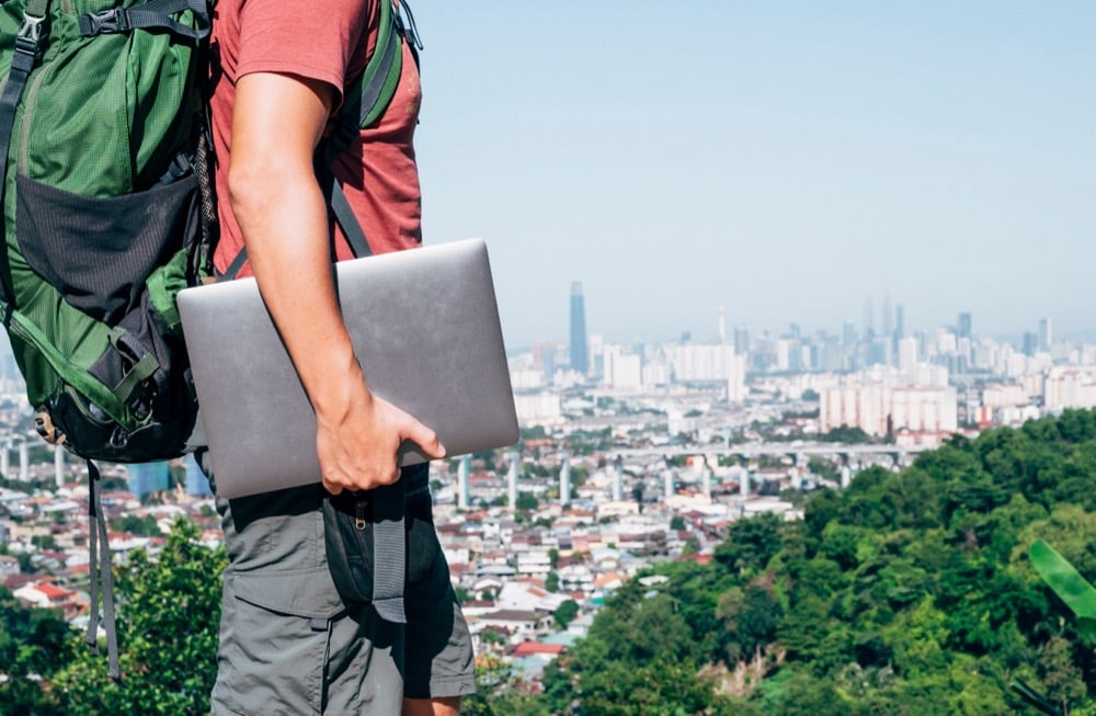 A digital nomad with a hiking backpack holds a laptop on a hilltop, overlooking a sprawling city skyline