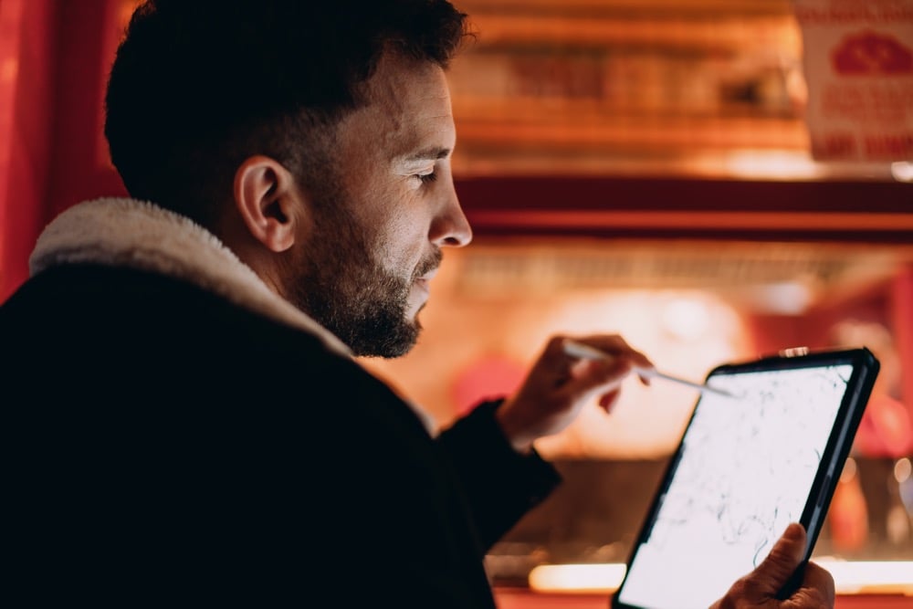 A bearded man drawing on a digital tablet with a stylus in a warmly lit restaurant setting
