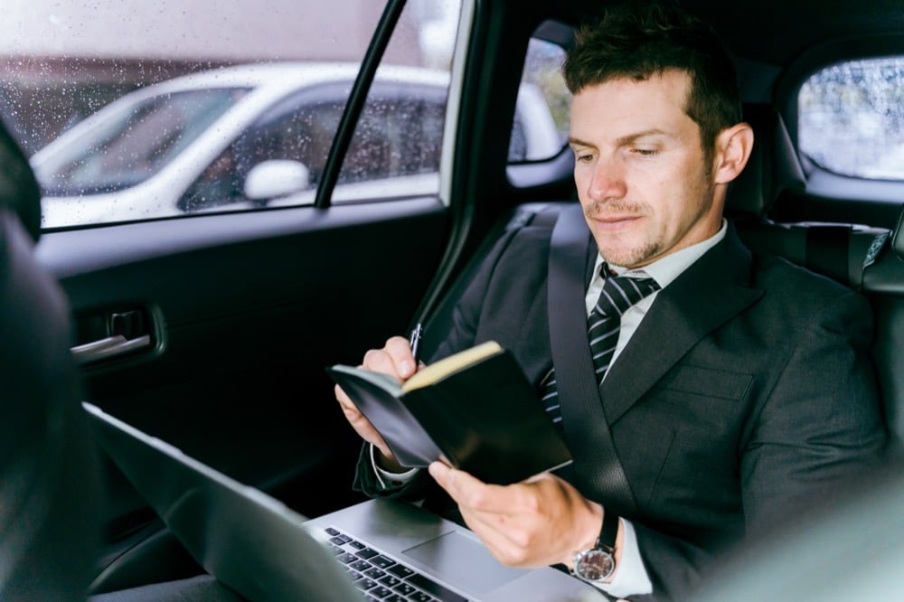 A suited businessman writes in a notebook with a laptop on his lap in a car's back seat