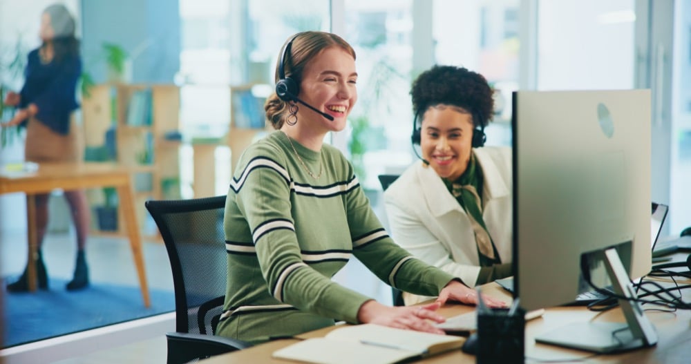 Two smiling women wearing headsets work at computers in a bright office