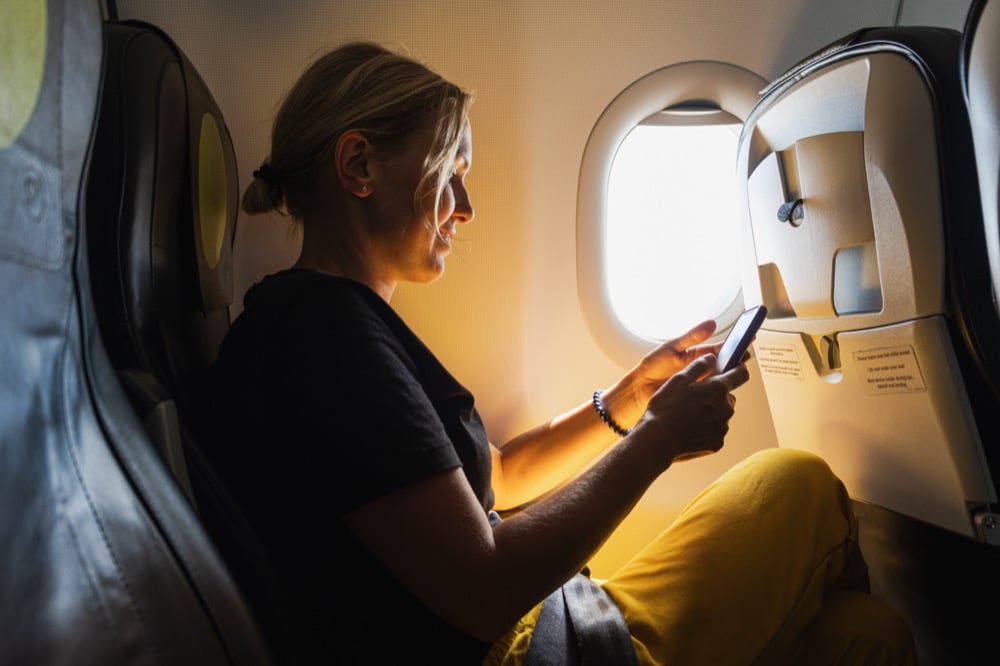 Woman sitting by an airplane window using a smartphone in the sunlight
