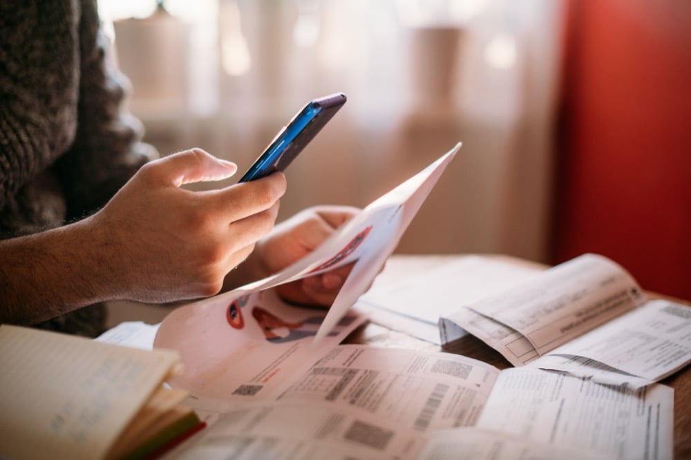 A person using a smartphone while sorting through a pile of bills and documents