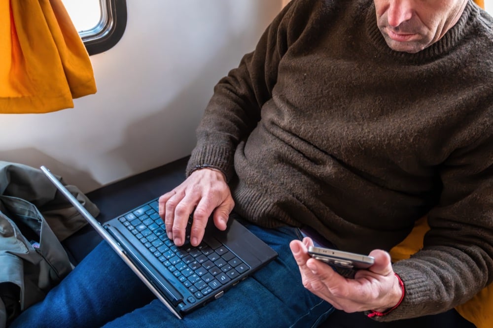 Man in a brown sweater using a smartphone and laptop on a train
