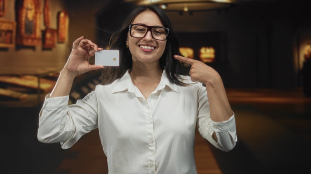A smiling woman with glasses pointing at a blank white credit card in a museum setting
