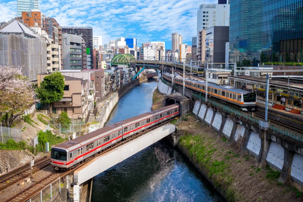 Multiple Tokyo metro trains crossing a river canal bridge surrounded by city buildings and cherry blossoms