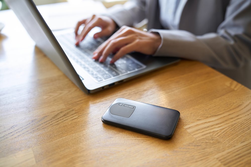 A smartphone wifi resting on a wooden desk with a person typing on a laptop nearby