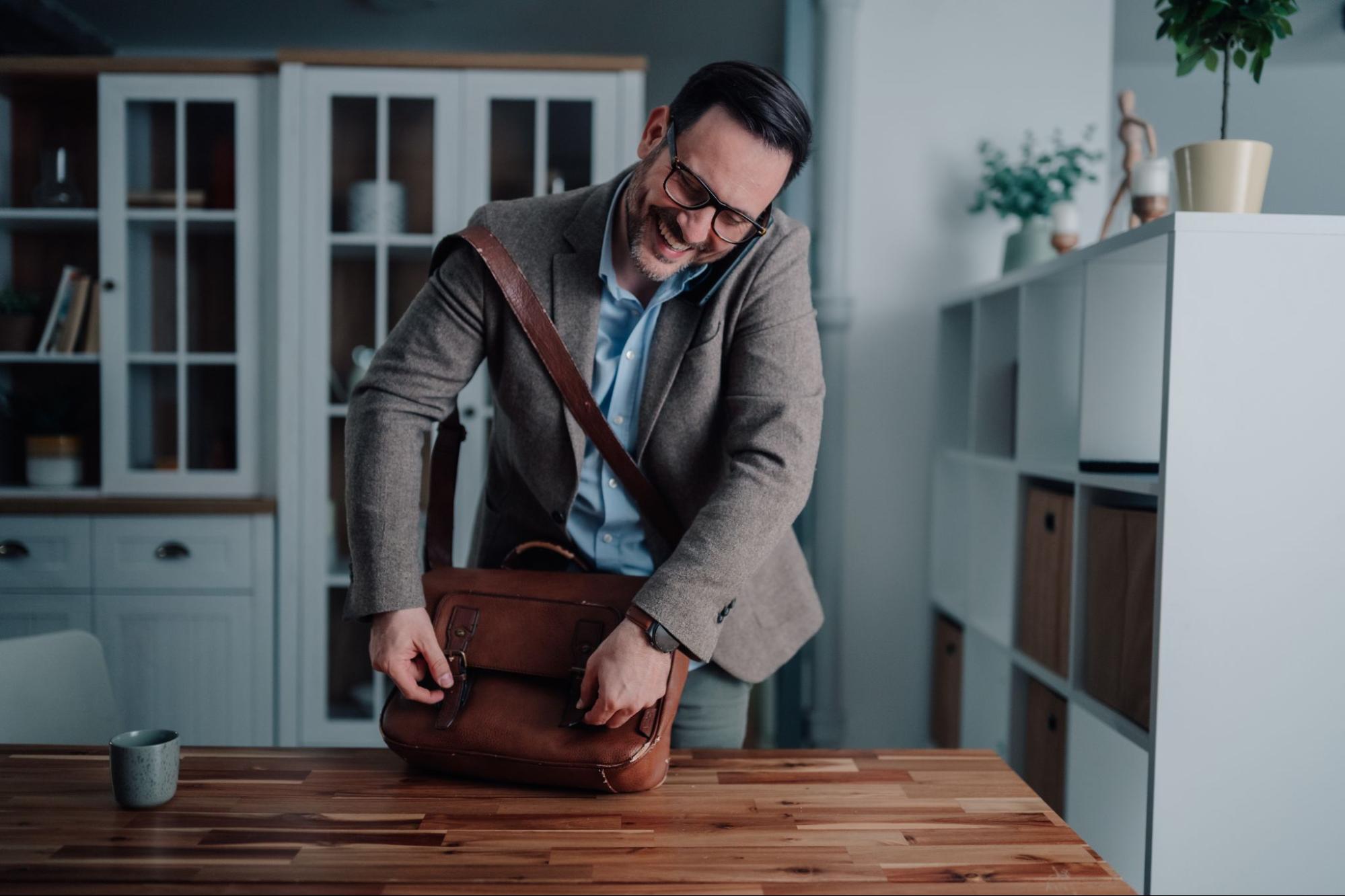 Smiling businessman in glasses talking on phone while opening a leather bag