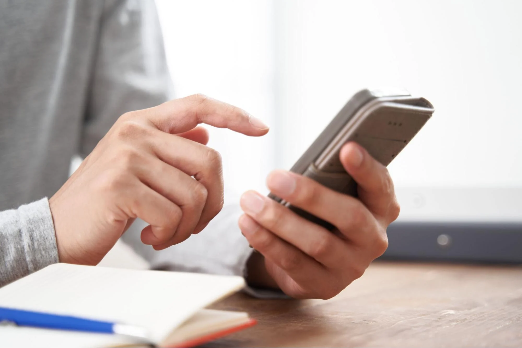 Close-up of hands tapping a smartphone screen on a desk with a notebook