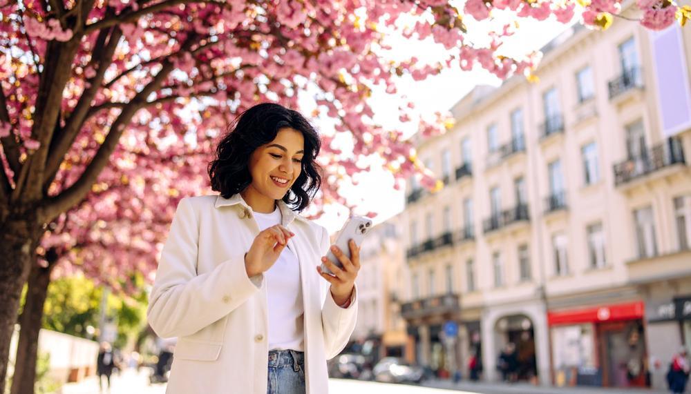 A woman is looking at her smartphone by pink cherry blossoms on a city street