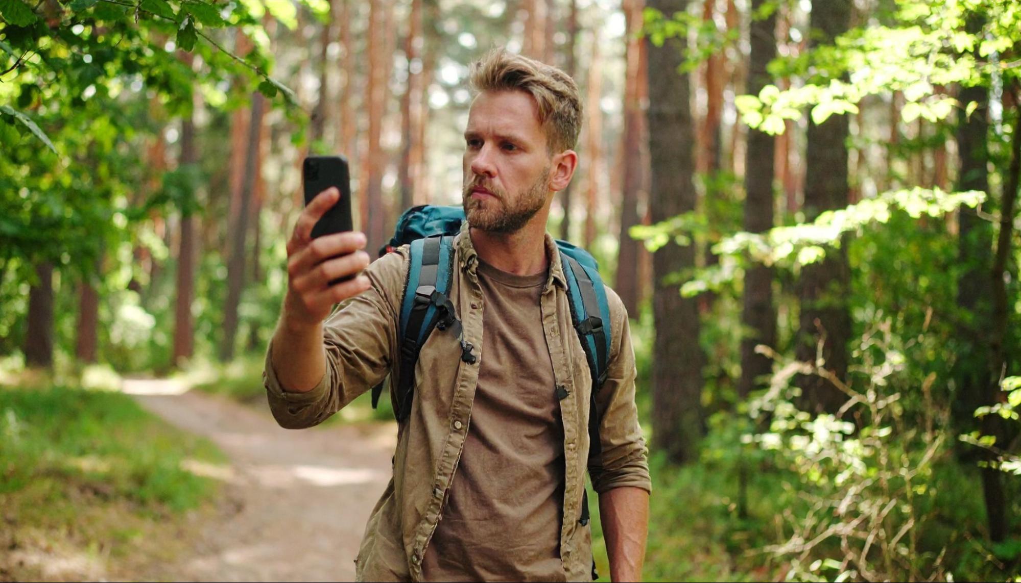 A man walks along a forest path, holding his smartphone up for navigation
