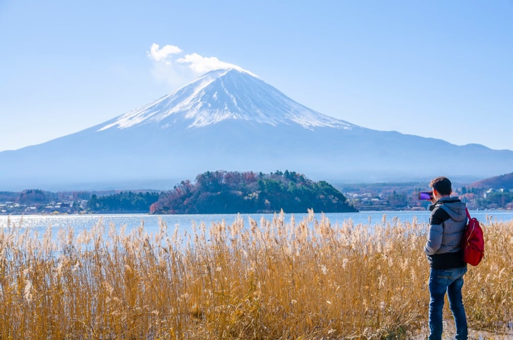 A man takes smartphone photo of Mount Fuji across a lake
