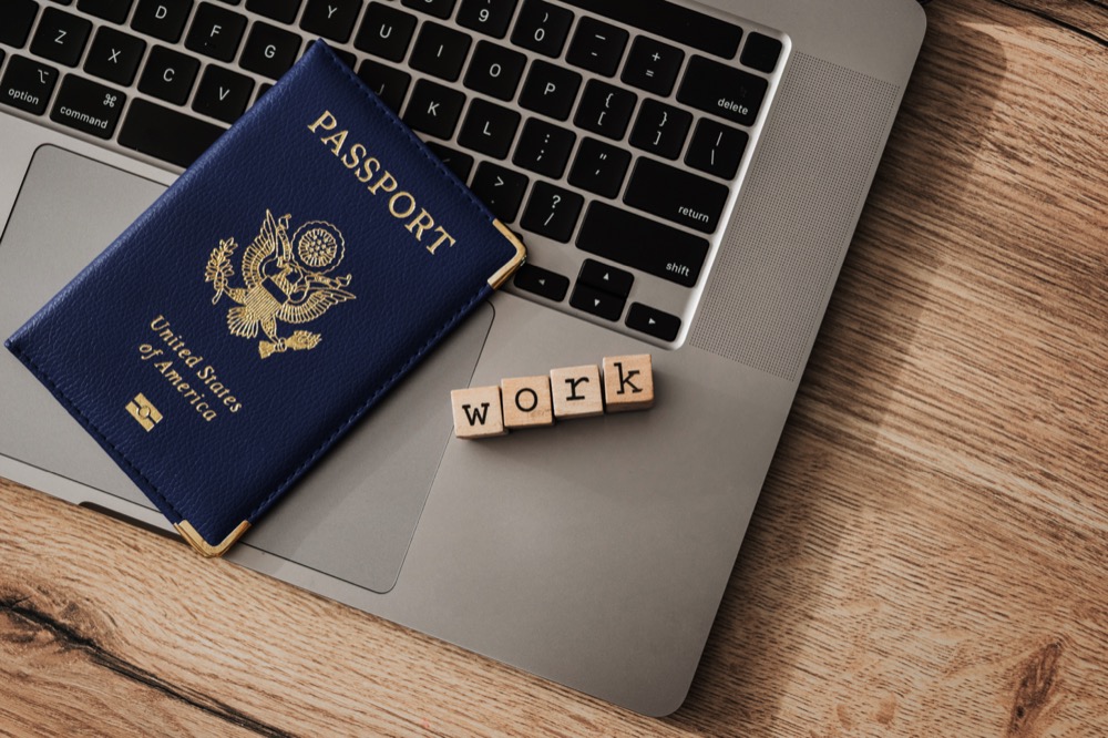 A US passport and wooden blocks spelling "work" rest on a MacBook, symbolizing remote work and travel