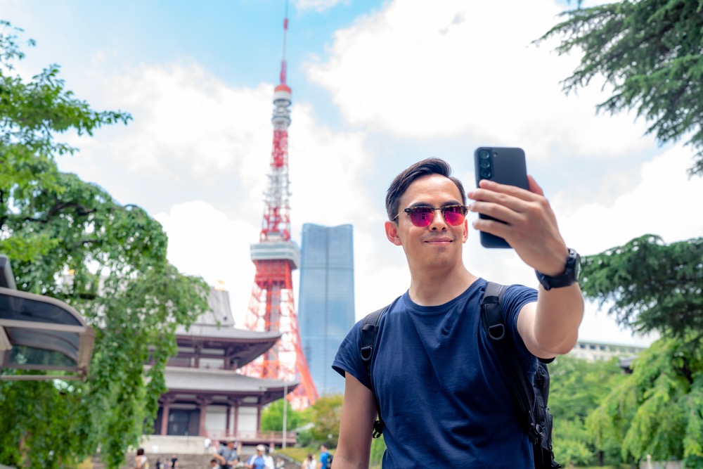A young male tourist taking a selfie in front of Tokyo Tower with a backpack and sunglasses