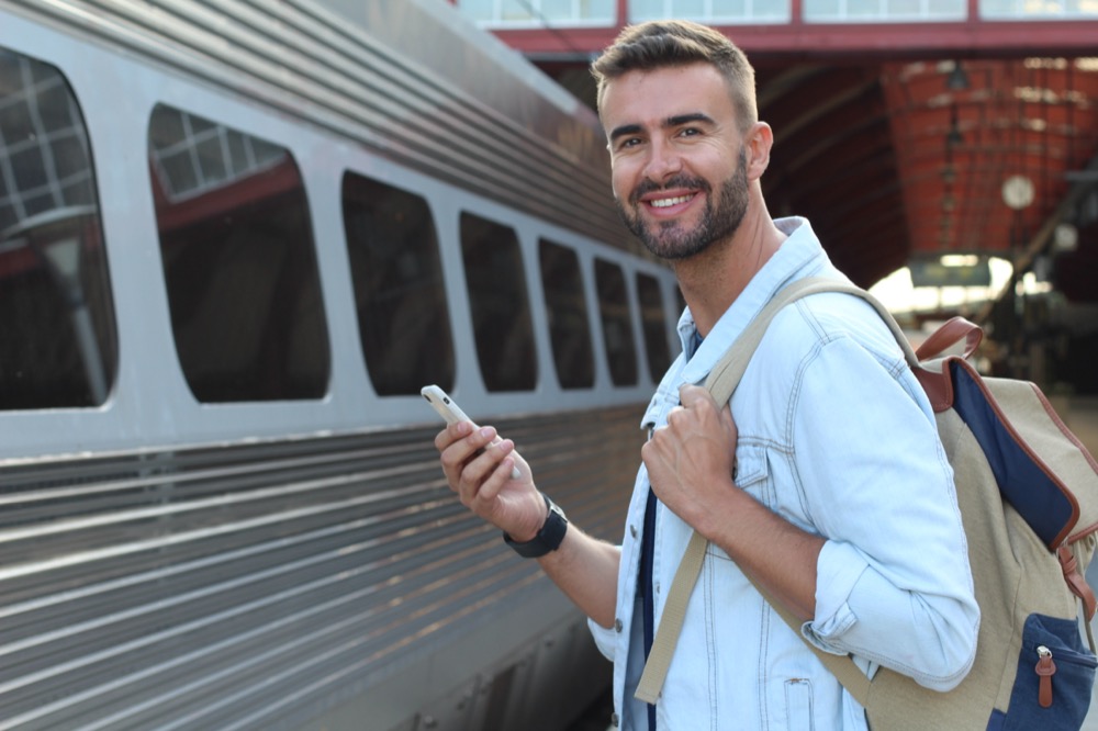 A smiling traveler with a backpack using a smartphone at a train station